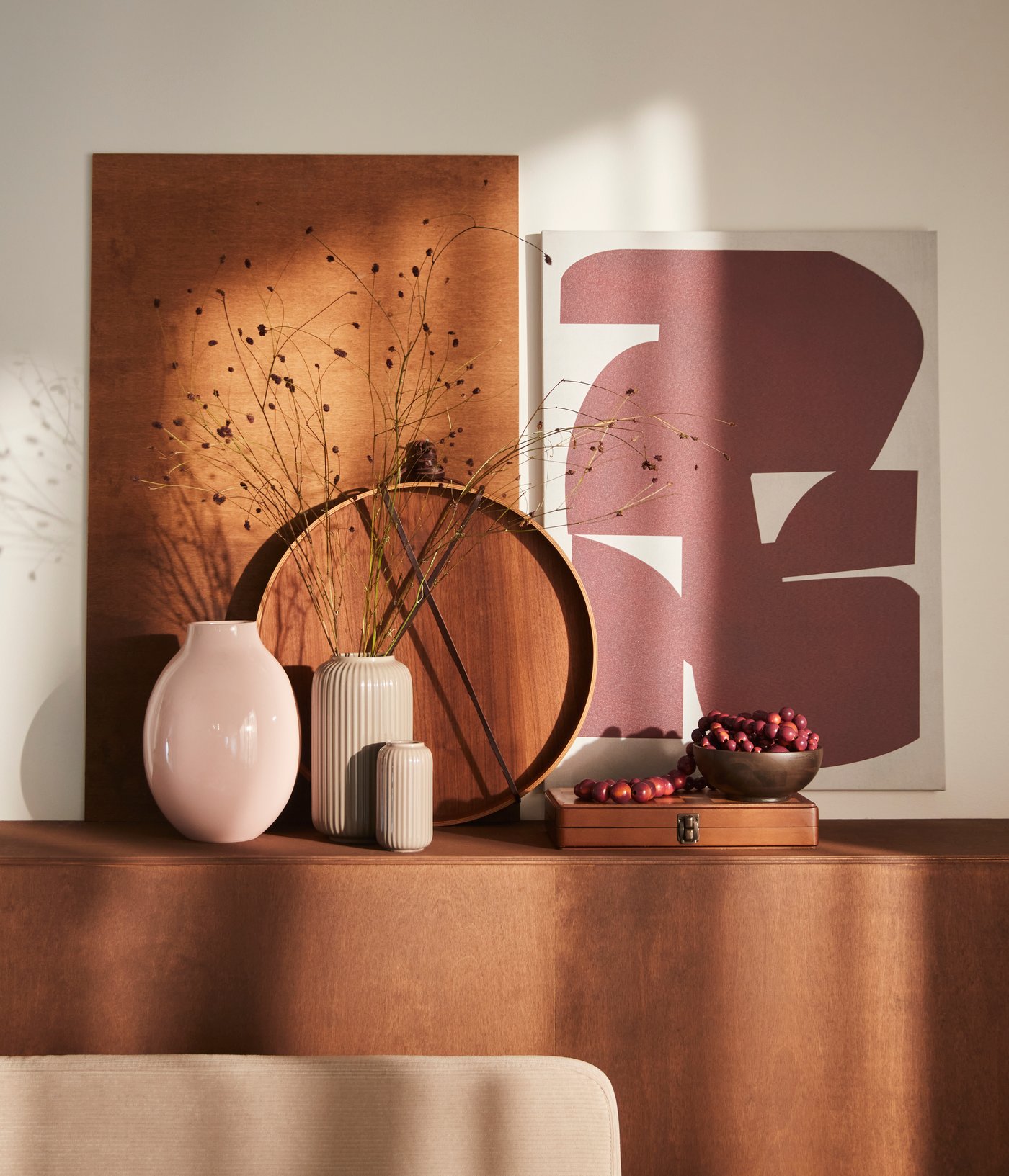 En sart pink BLODBJÖRK vase står på en gyldenbrun overflade sammen med flere vaser, dekorative genstande og et PJÄTTERYD billede.