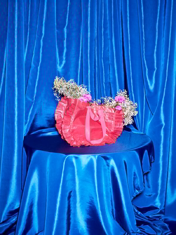 A medium KARISMATISK pink carrier bag sits atop a table completely covered in blue satin; the bag is filled with flowers.