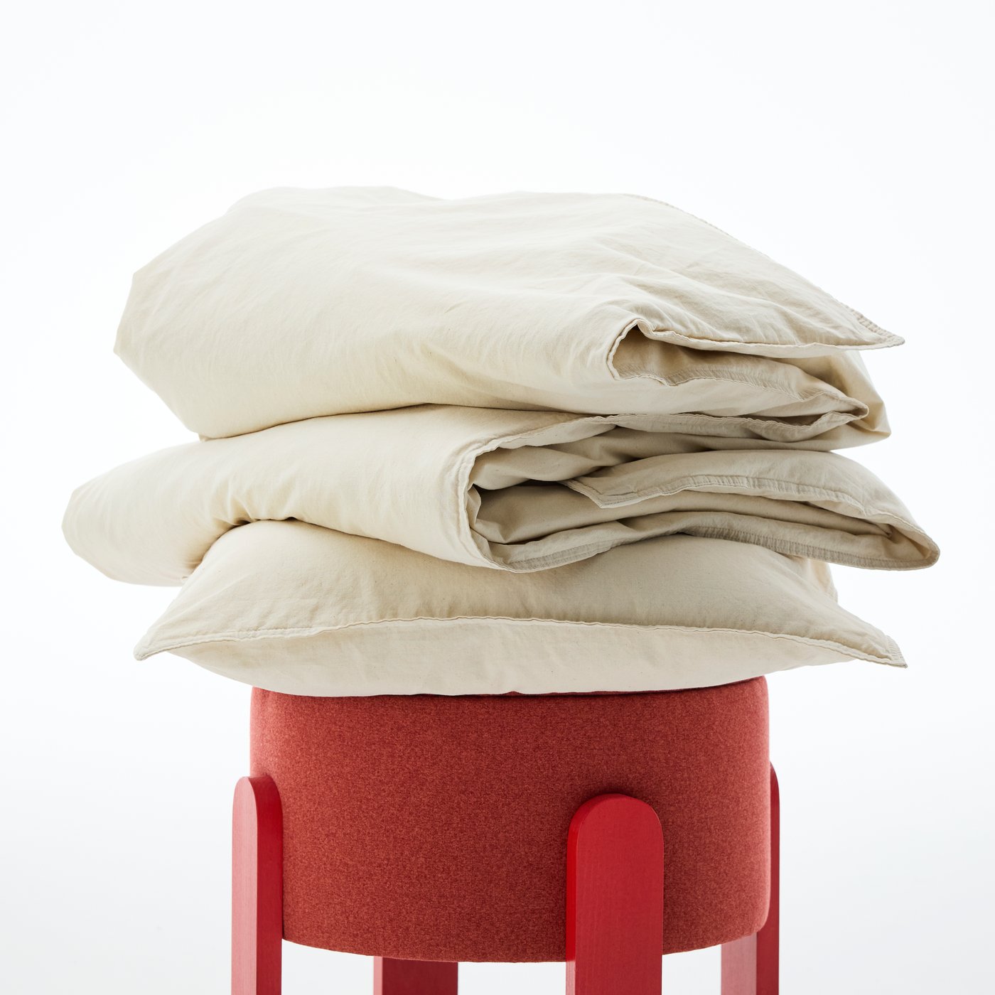 Close-up of a natural cotton colour set of ÄNGSLILJA duvet and pillowcase, piled on top of a red stool.