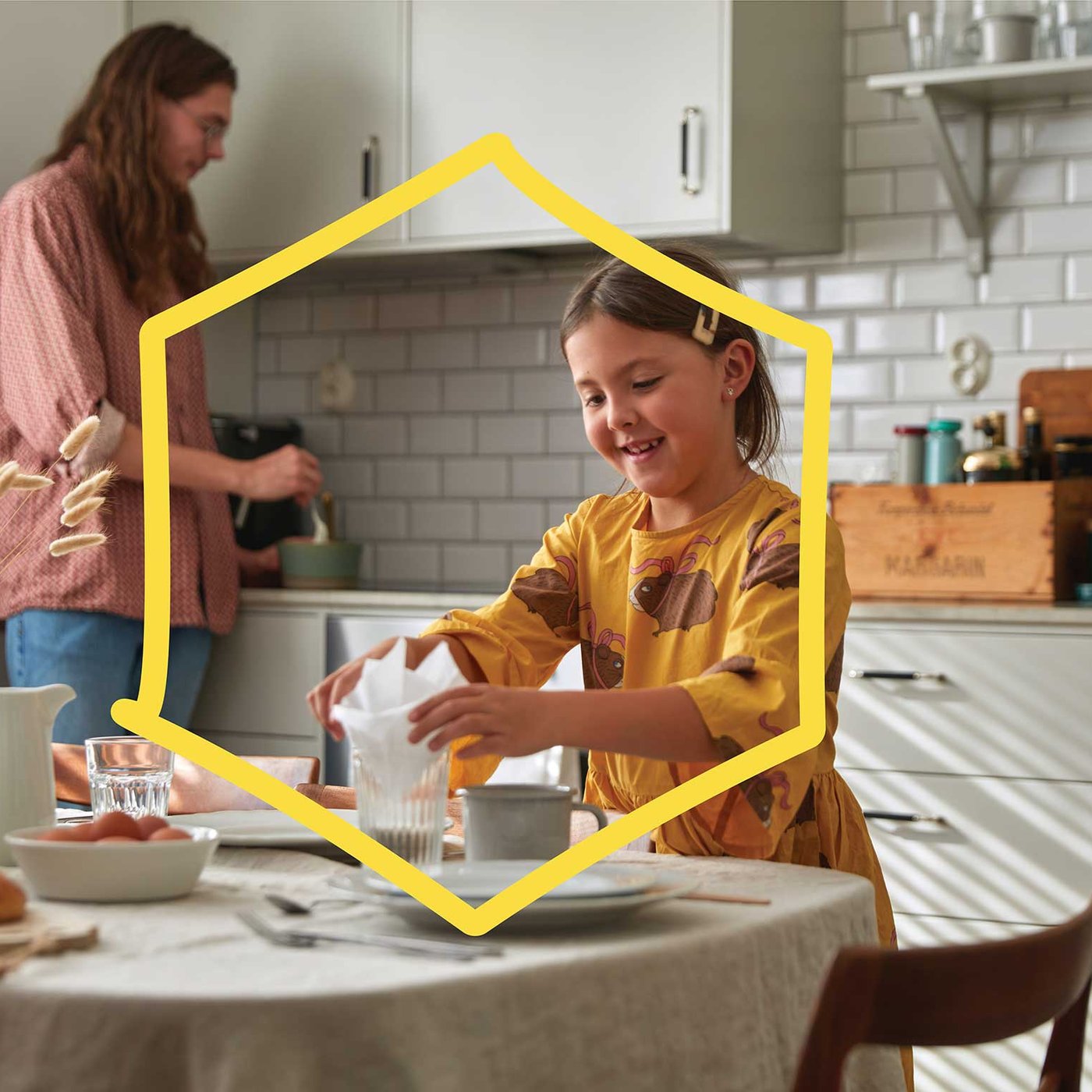 Un enfant vêtu d'une robe jaune place une serviette dans un verre à table, tandis qu'une autre personne remue des ingrédients dans un bol dans une cuisine lumineuse aux carreaux de sol blancs.
