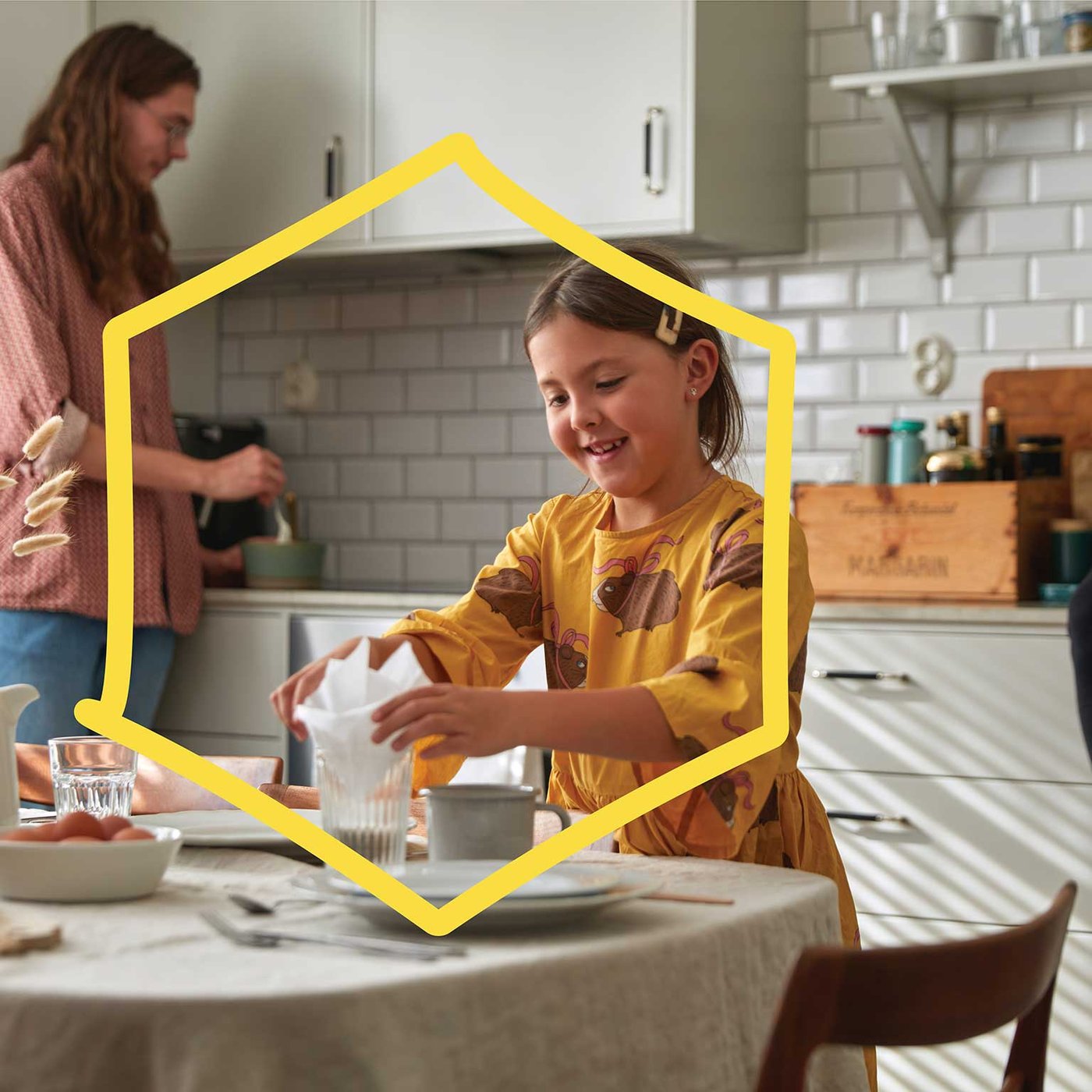 Een kind in een gele jurk legt een servet in een glas aan tafel, terwijl een andere persoon ingrediënten in een kom roert in een lichte keuken met witte metrotegels.