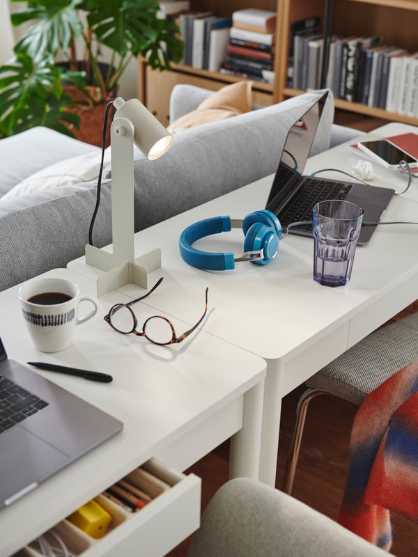 Two white TONSTAD desks are placed next to each other behind a sofa. There are two laptops and a cup of coffee on the desks.