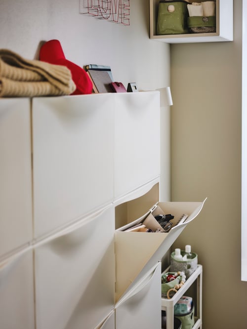 A side view of a white TRONES shoe cabinet with one compartment drawn open. A VESKEN trolley is next to the cabinet.