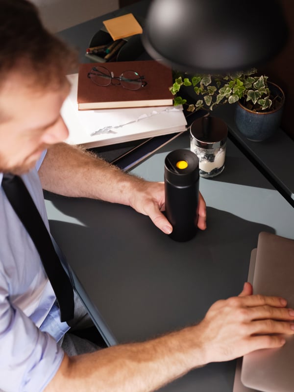 A man sitting at his desk, opening his laptop with one hand and holding a black UNDERSÖKA insulated travel mug in the other.