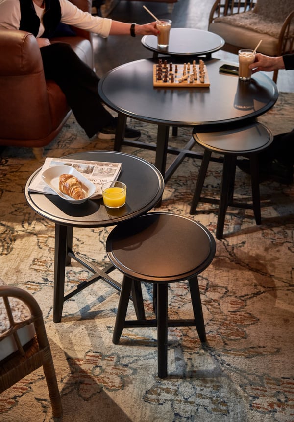 Flera svarta bord med fika och schackbräde, två personer delvis synliga, mönstrad matta och mysig inomhusmiljö.