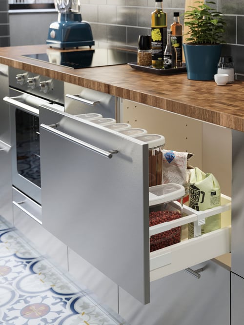 A kitchen has a wooden worktop on METOD/MAXIMERA base cabinets with stainless steel drawer fronts. An open one has dry goods.