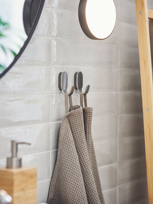 Two beige hand towels are hanging on two BROGRUND hooks made of stainless steel are mounted on a grey-tiled bathroom wall.