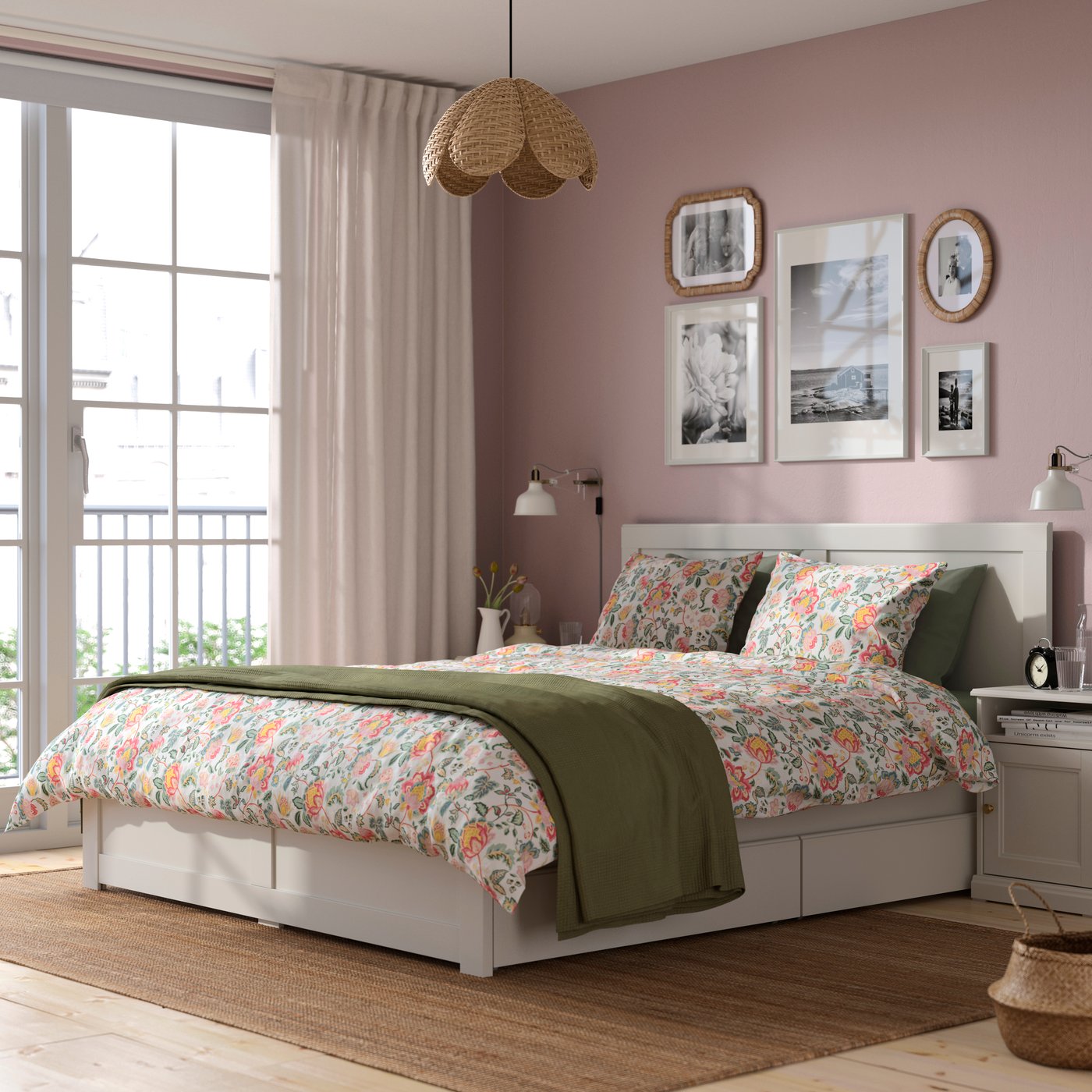 A stylish bedroom with a floral-patterned bed, blush pink walls, framed artwork, and soft natural light from a large window.