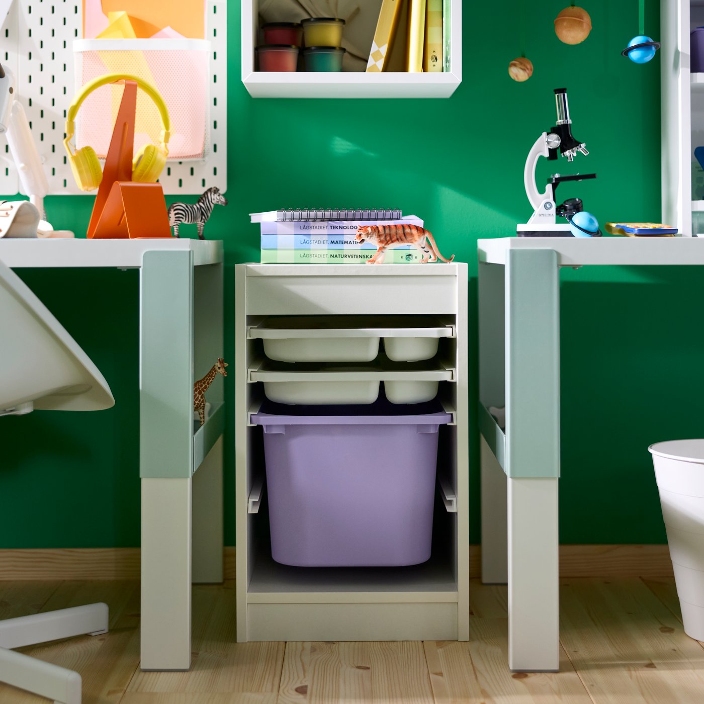 A TROFAST storage combination with trays and a large box placed between two PAHL desk legs in a children’s study room.
