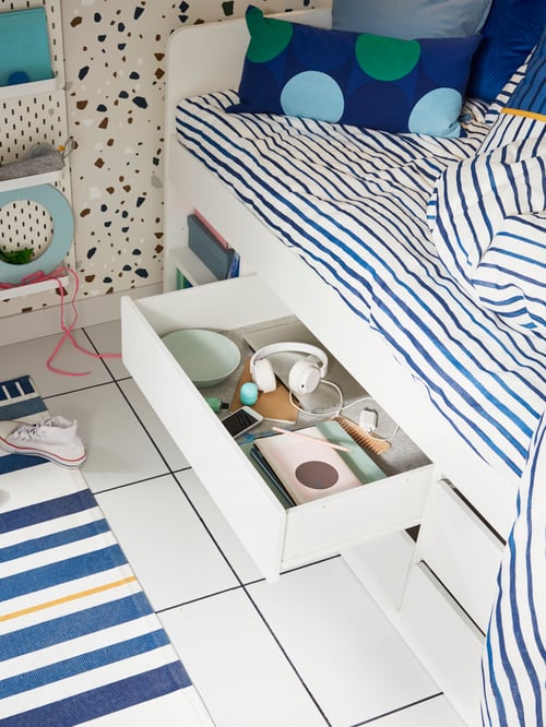 White SLÄKT bed with an opened storage drawer to show ear phones and various items, striped bed linen and coloured pillows.