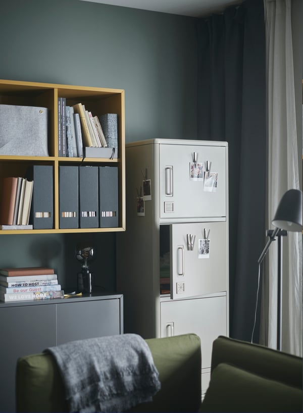 IDÅSEN beige high cabinet stands by a wall. Magnetic clips with photos are put on some of the cabinet doors.