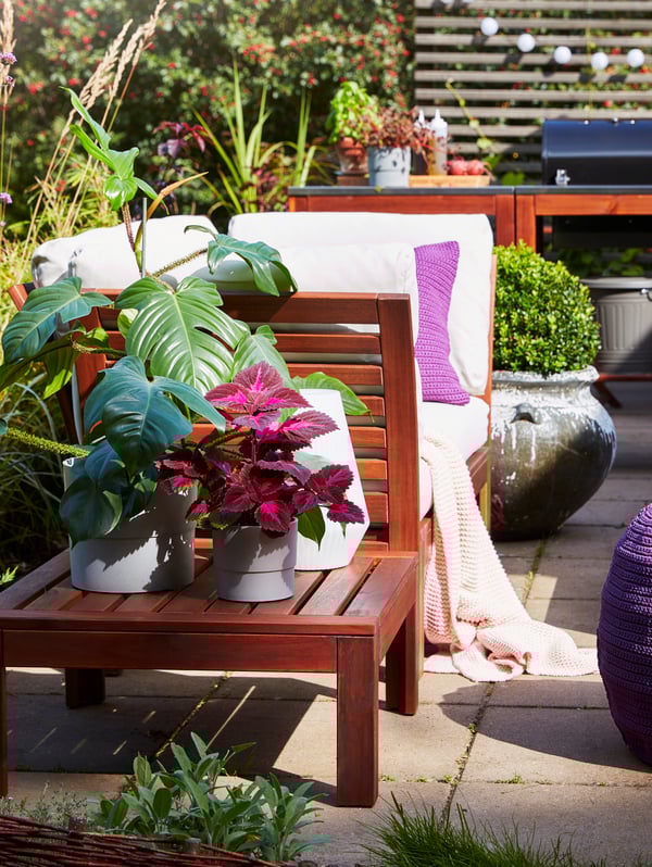 A brown-stained table with plants in grey plant pots stands next to an outdoor sofa with cushions and a throw.