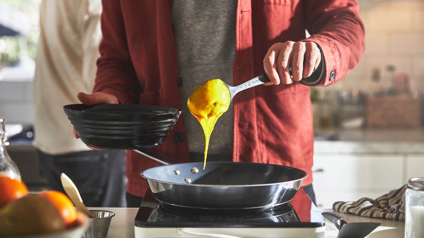 En person i röd jacka häller gul smet från en sked till en stekpanna. Personen håller även en svart skål. I förgrunden syns frukt på en köksbänk.