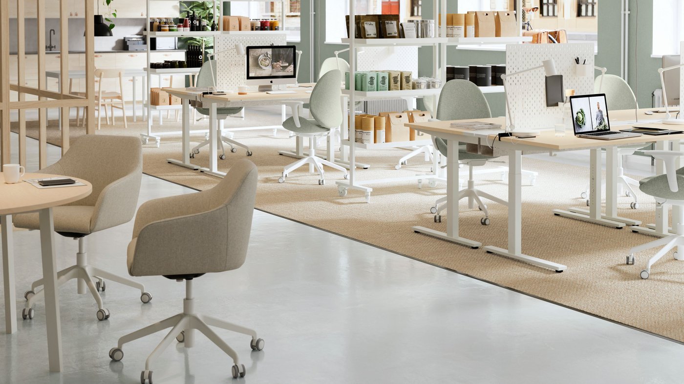 Ett modernt och ljust kontorslandskap med flera arbetsstationer utspridda i ett öppet rum. Skrivborden är ljusa och höj‑ och sänkbara, och vid dem står stolar med mjuk, ljus klädsel på hjul. På borden finns laptops och några mindre kontorsdetaljer. I bakgrunden syns hyllor fyllda med lådor, böcker och förvaring. Golvet består av en kombination av slätt, ljusgrått material och en stor beige matta som markerar arbetszonerna. Rummet har stora fönster som släpper in mycket naturligt ljus och skapar en öppen, rymlig känsla.