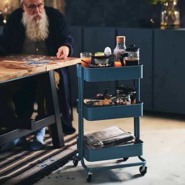 An elderly man with a long beard lays a jigsaw puzzle on his dining table. Beside him is a RÅSKOG trolley with his breakfast.