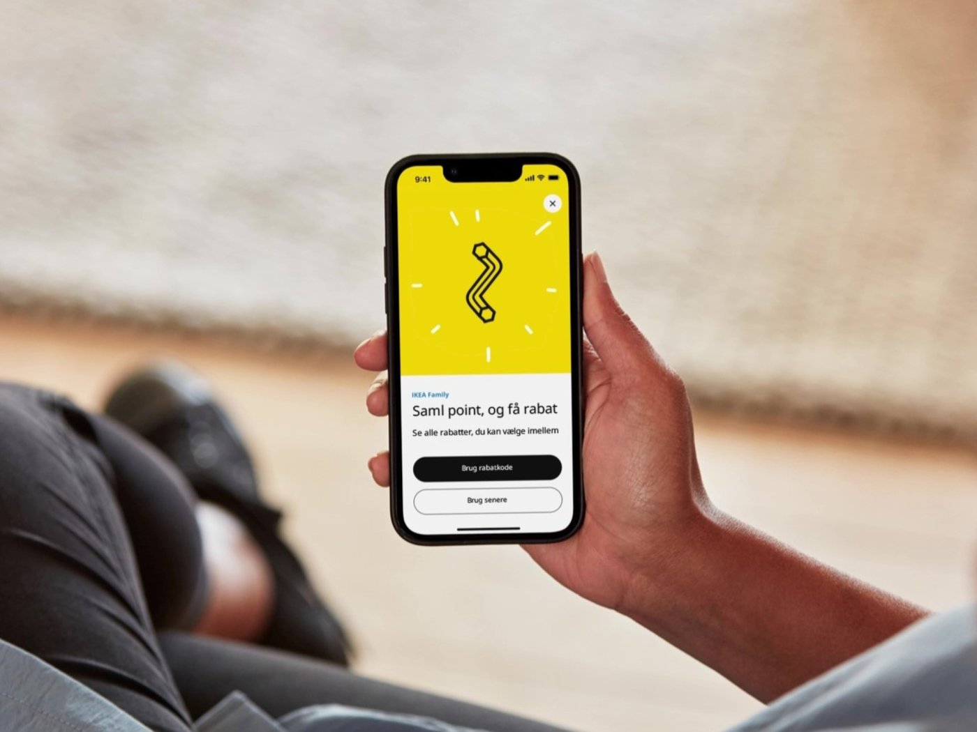 En hånd holder en telefon og på skæren kan ses en unbracho nøgle på en gul baggrund og der står at du som IKEA Family medlem kan samle point.