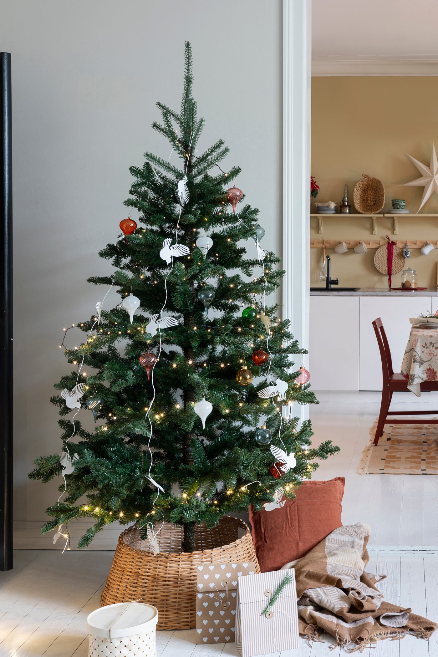 En grønn VINTERFINT kunstig juletre i stuen dekorert med julekuler og en VINTERFINT juletrefotskjuler i rotting.