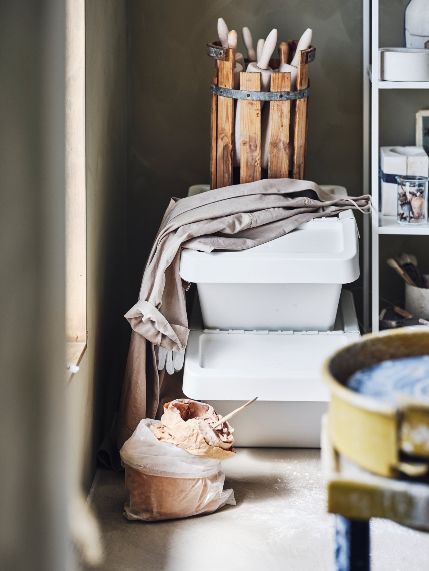 Espacio de trabajo con cubos de reciclaje blancos con tapa, una estantería blanca, ropa, productos de limpieza, detergentes y ropa sucia.