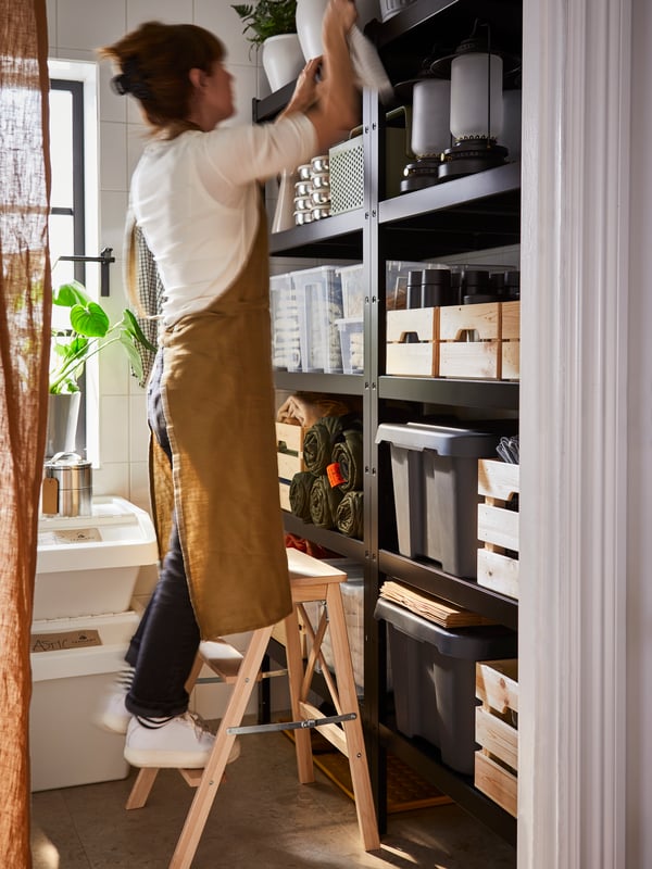 A person wearing a white t-shirt, black jeans and an apron is standing on a BEKVÄM stepladder next to a BROR shelving unit.