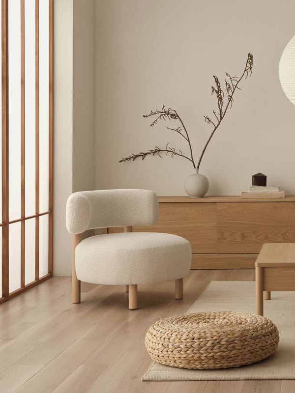 Minimalistiskt vardagsrum i Japandi-stil med ljusa trägolv, krämfärgad stol, träbord, puff och sideboard med vas och torkade grenar.