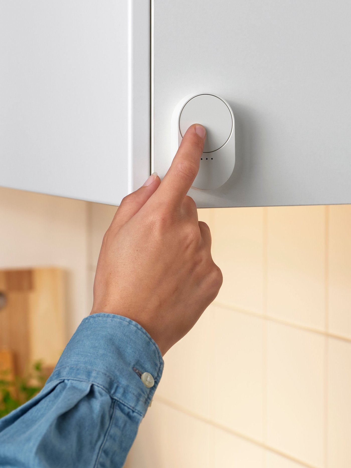Someone is touching a white smart BILRESA remote control with a scroll wheel that’s attached to a kitchen cabinet.