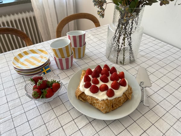 På ett bord med rutig duk syns en tallrik med en pannkakstårta, färgglatt randigt porslin, en skål med jordgubbar och en vas med blommor i.