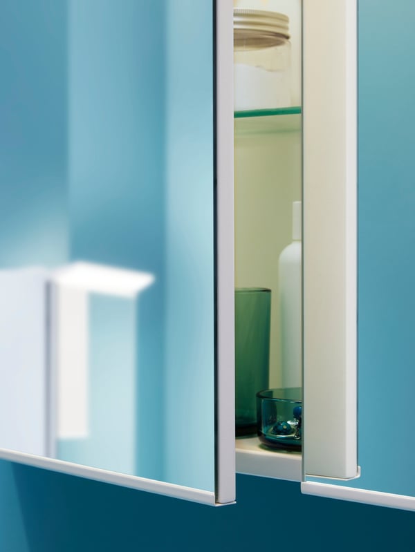 A TREASJÖN mirror cabinet on a teal-colored wall, with one of cabinet doors slightly opened, revealing a toothbrush holder.