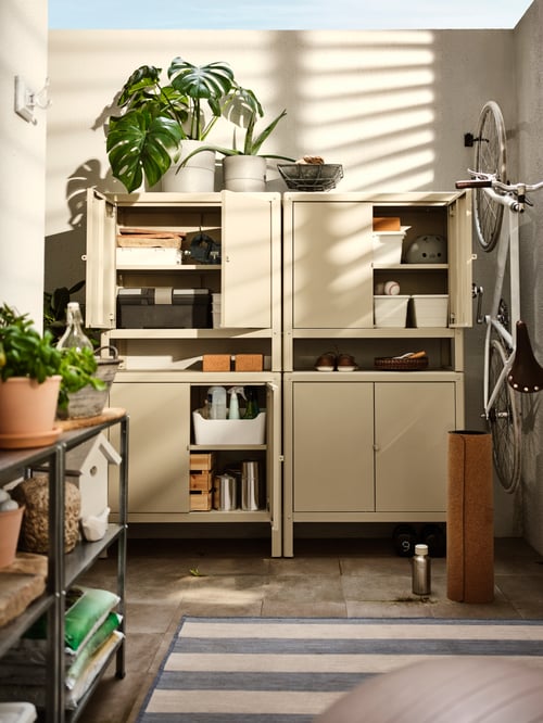 Four stacked beige KOLBJÖRN cabinets with four doors open showing items inside, plus various plants and a hanging bicycle.