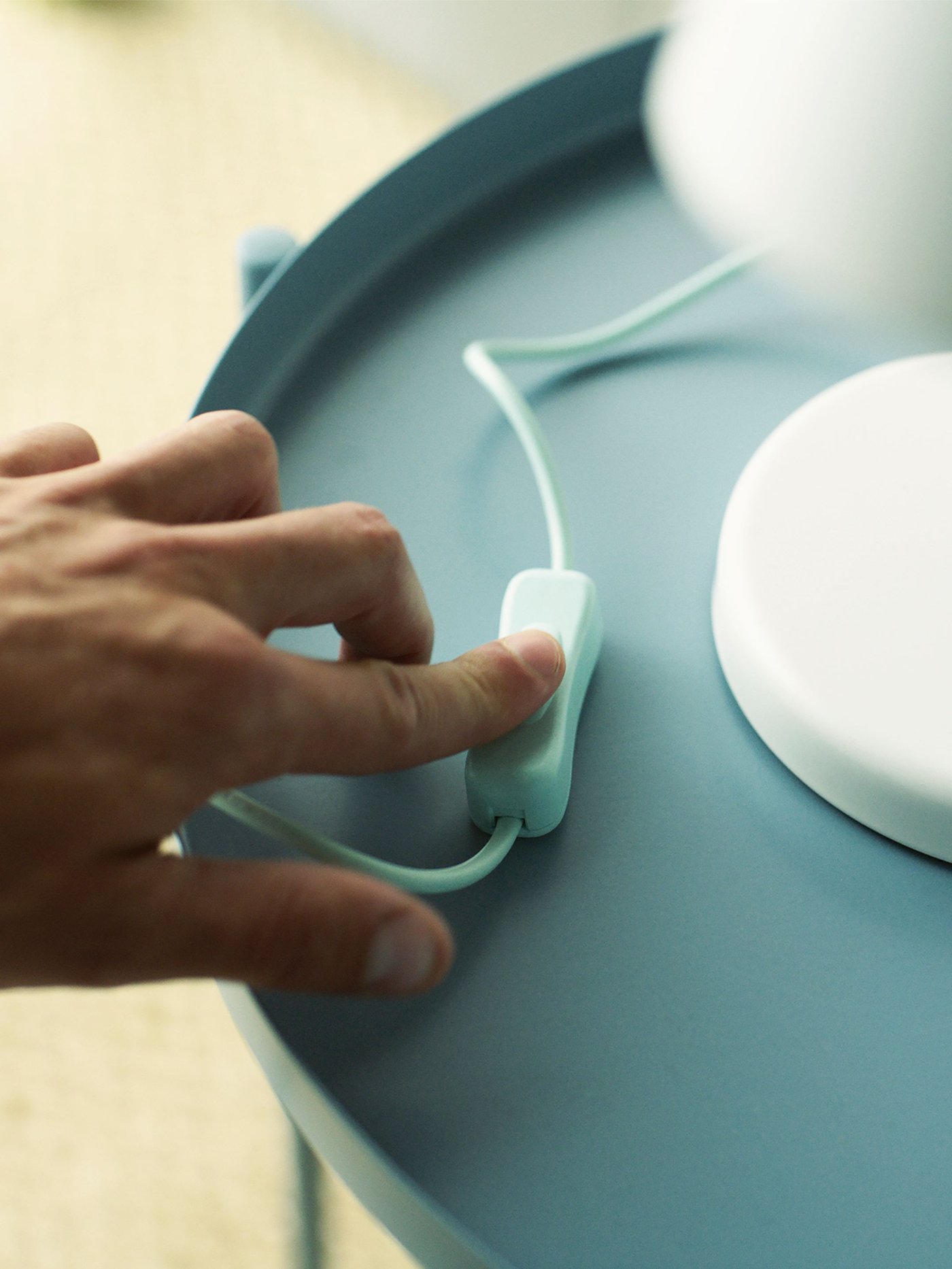 Dit d'una persona que pressiona l'interruptor d'encesa/apagada del cable d'un llum de treball LED FUBBLA de color blanc sobre una taula amb safata de blau clar.