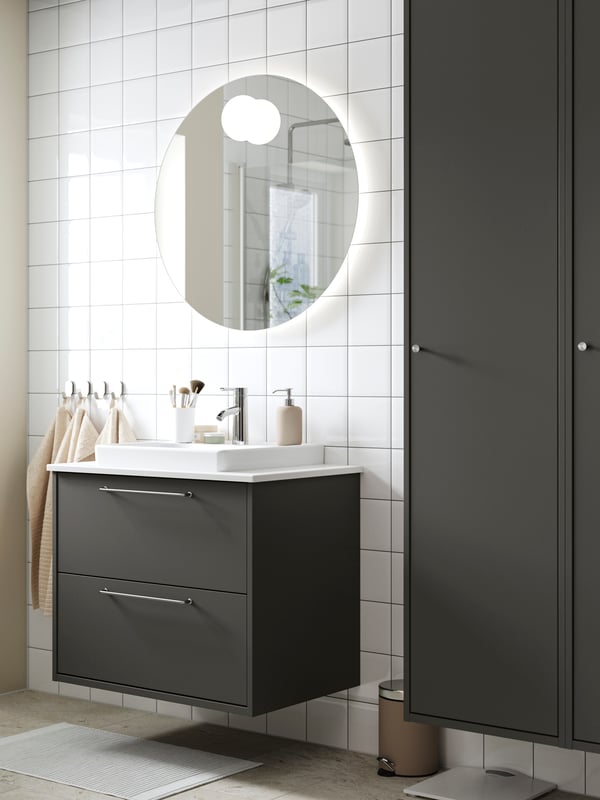 Un meuble lavabo HAVBÄCK gris foncé à tiroirs est fixé sur un mur carrelé blanc avec un miroir rond avec éclairage intégré.