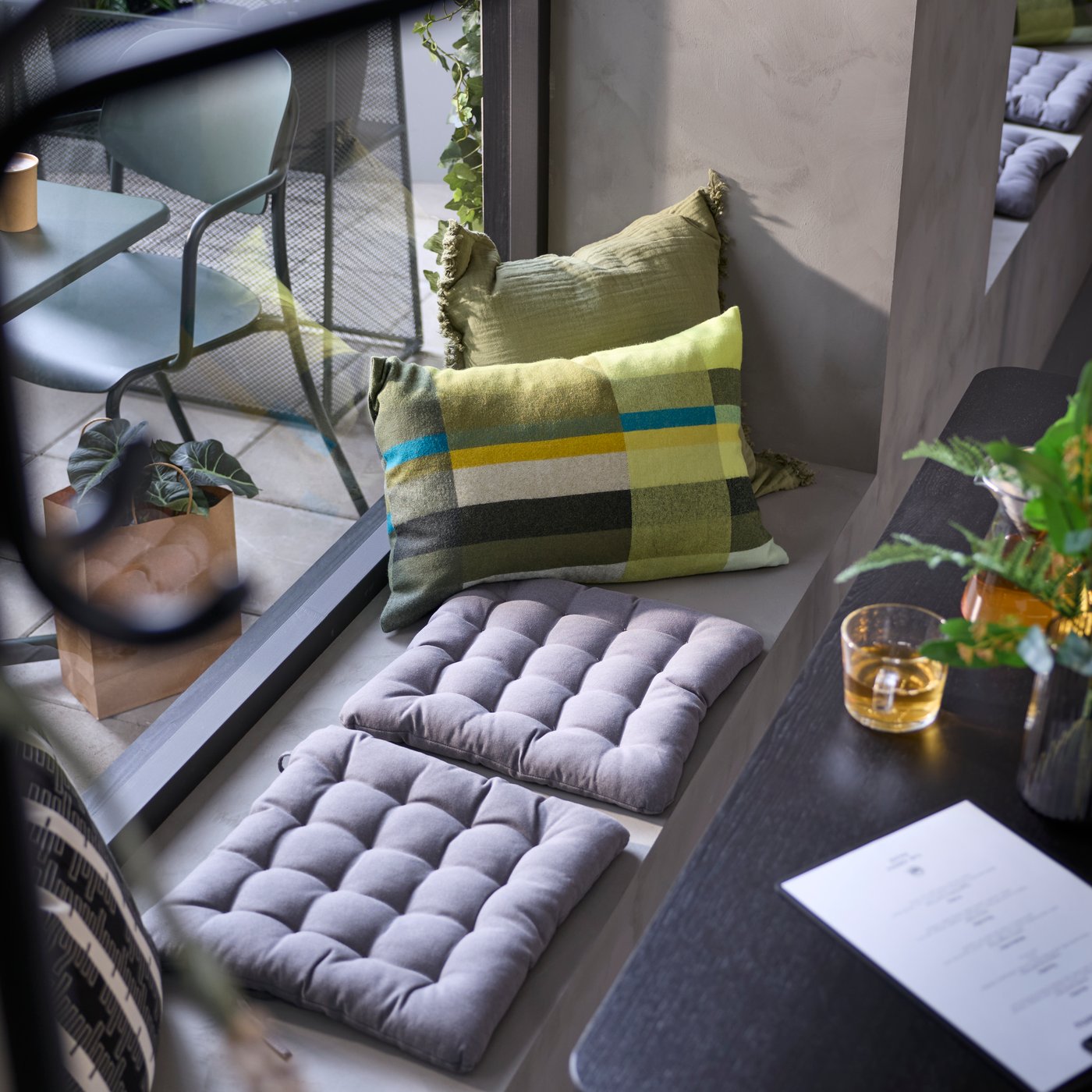 A sunlit corner with dark grey VIPPÄRT chair pads on a grey seating area, surrounded by green plants, pillows and a table.