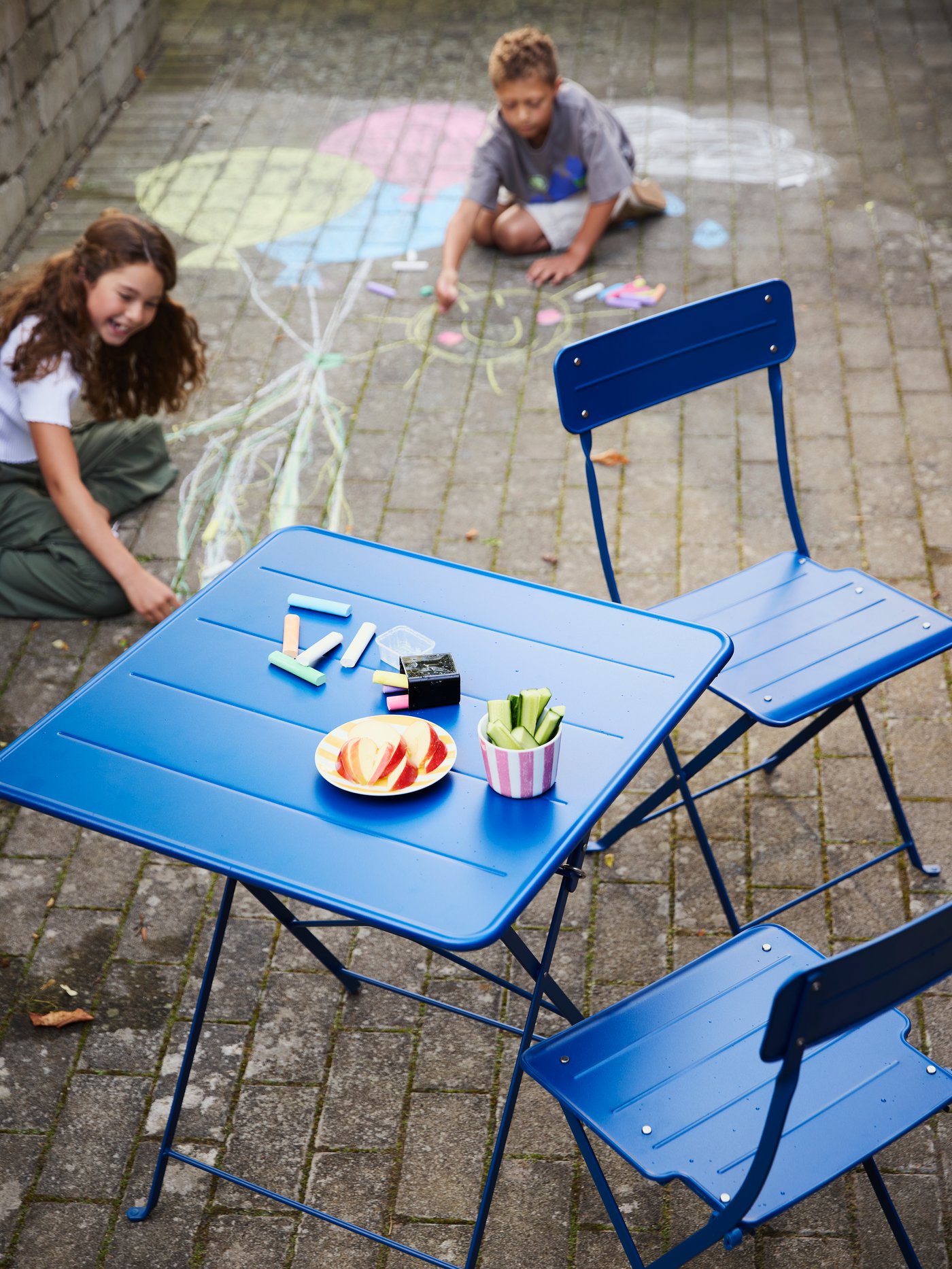Tavolo SUNDSÖ blu in un cortile, con due bambini sullo sfondo che disegnano sul pavimento con i gessetti MÅLA.