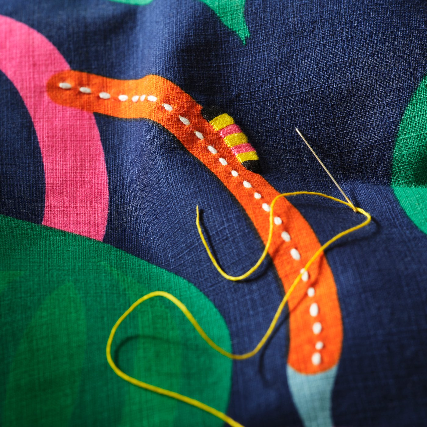A close-up of the embroidered caterpillar on the MÄVINN cushion cover in dark blue. On top lies a needle with yellow thread.