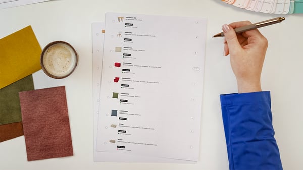 Projektantka wnętrz IKEA prezentująca kartki z wydrukiem listy produktów wykorzystanych w zaprojektowanym pomieszczeniu. Obok na blacie leży wzornik kolorów i próbki materiałów.