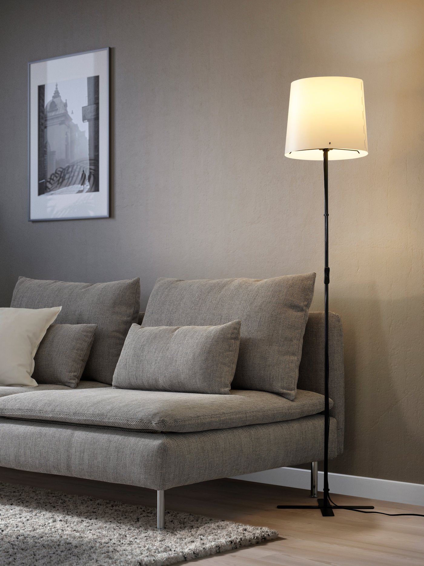 Een zwart/witte BARLAST vloerlamp naast een grijze bank in de woonkamer.