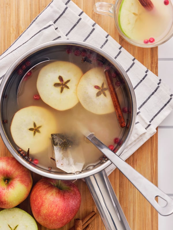 A saucepan of FLÖJT green tea with sliced apples, cinnamon sticks, and spices is placed on a tea towel on a wooden surface.