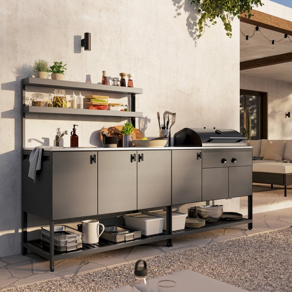 A dark grey BÅTSKÄR outdoor kitchen with charcoal barbecue. Above the closed cabinets are two open shelves with fresh herbs.