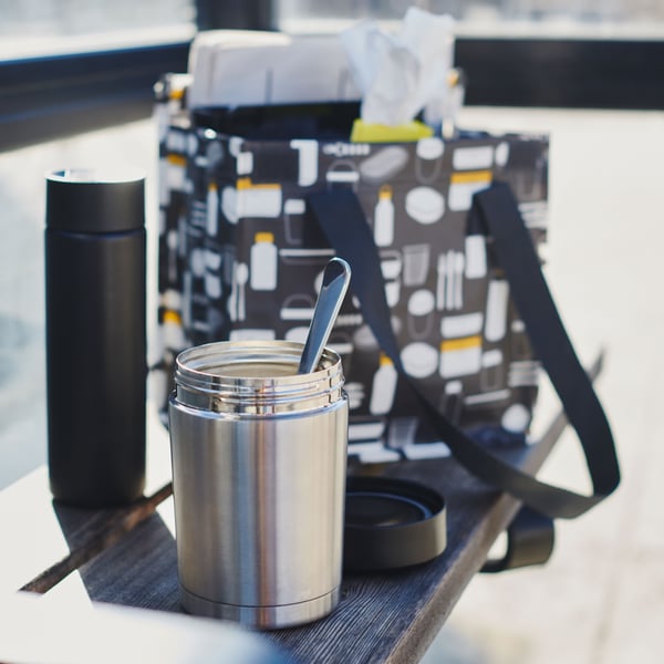 En öppen EFTERFRÅGAD termos med en sked i. Placerad framför en FLADDRIG matkasse på en solig bänk.