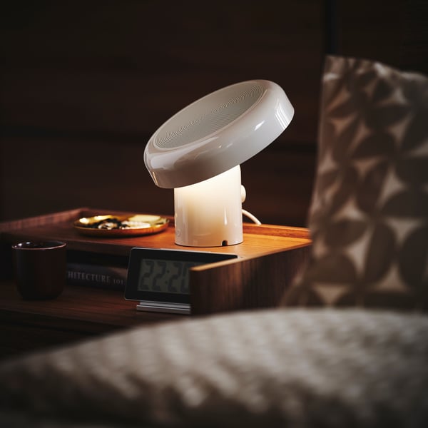 Beige BLOMPRAKT-kaiutinvalaisin yöpöydän päällä pimeässä huoneessa.