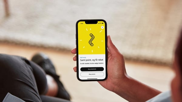 En hånd holder en telefon, hvor der er et billede af en sort unbraco nøgle på en gul baggrund og nedenunder står der saml point og få rabat.