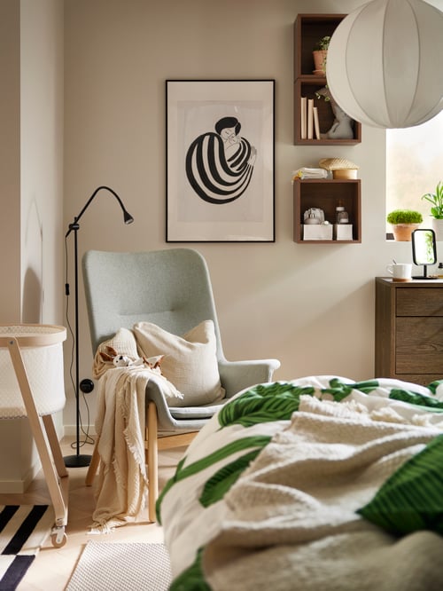 Schlafzimmer mit einem VEDBO Sessel mit hoher Rückenlehne neben einem beigefarbenen ÄLSKVÄRD Stubenwagen mit Matratze und einem Bild in einem LOMVIKEN Rahmen.