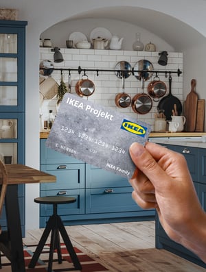 Hand holding an IKEA Projekt credit card in a modern kitchen with blue cabinets and hanging copper cookware.
