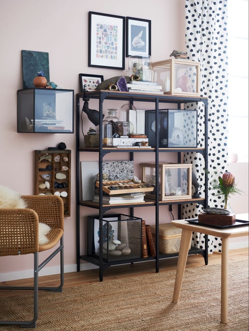 A FJÄLLBO shelving unit in black holds books and glass display boxes full of stones and geodes in a living room.
