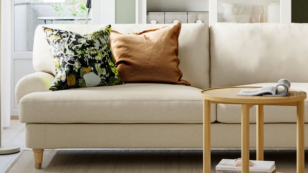 Ljusbeige VALNÄS soffa med kuddar I brunt och svart och ett runt soffbord i trä i ljust vardagsrum.