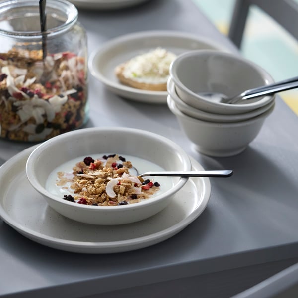 A light beige DAGGASTER deep plate holds granola and milk, placed next to some stacked DAGGASTER bowls and a jar of granola.