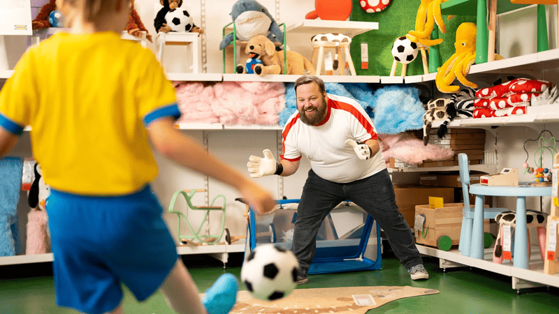 Eine Person mit einem Fußballtrikot versucht den Ball, den ein Kind in seine Richtung schießt, aufzufangen.