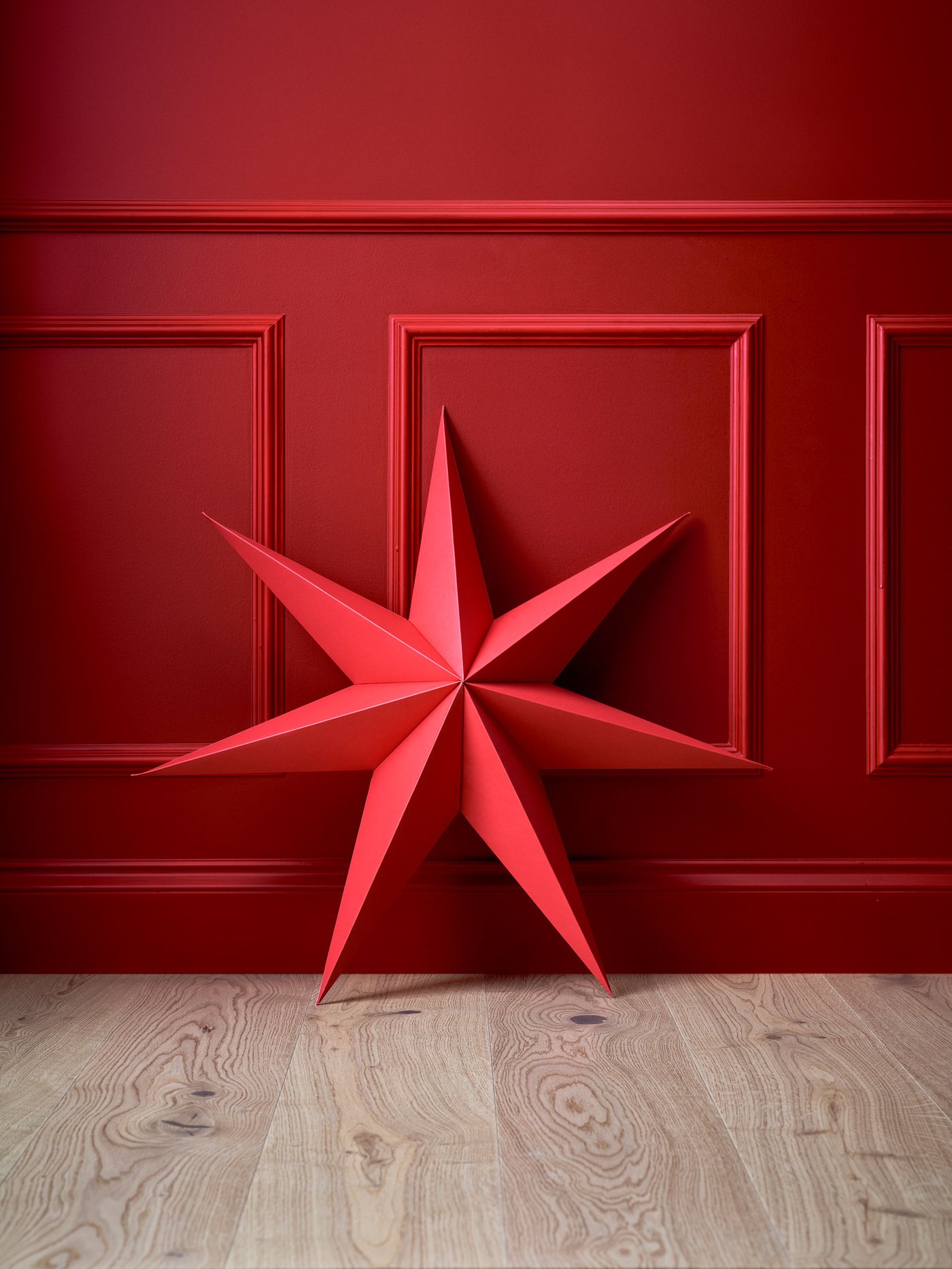 Stor röd adventsstjärna placerad mot en röd vägg på ett ljust trägolv.