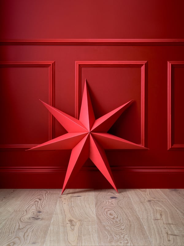 Stor röd adventsstjärna placerad mot en röd vägg på ett ljust trägolv.