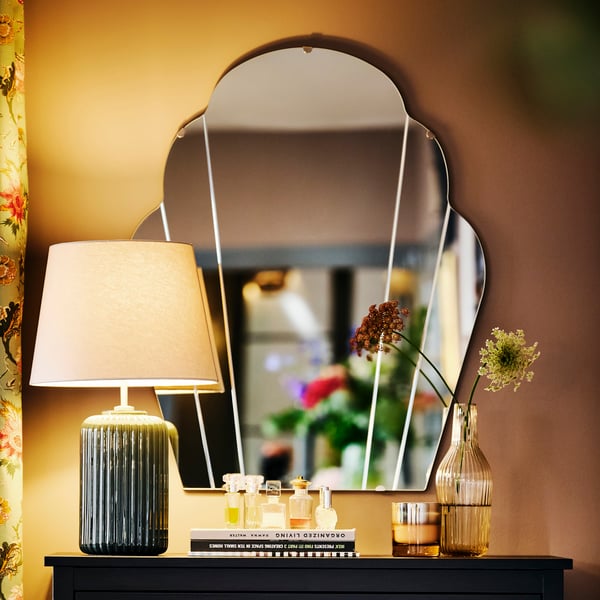 Dekorative Anrichte mit einem Spiegel, einer Lampe mit beigem Schirm und grünem Fuß, mehreren kleinen Flaschen und zwei Vasen mit Blumen, vor einer warmfarbenen Wand und floralen Vorhängen