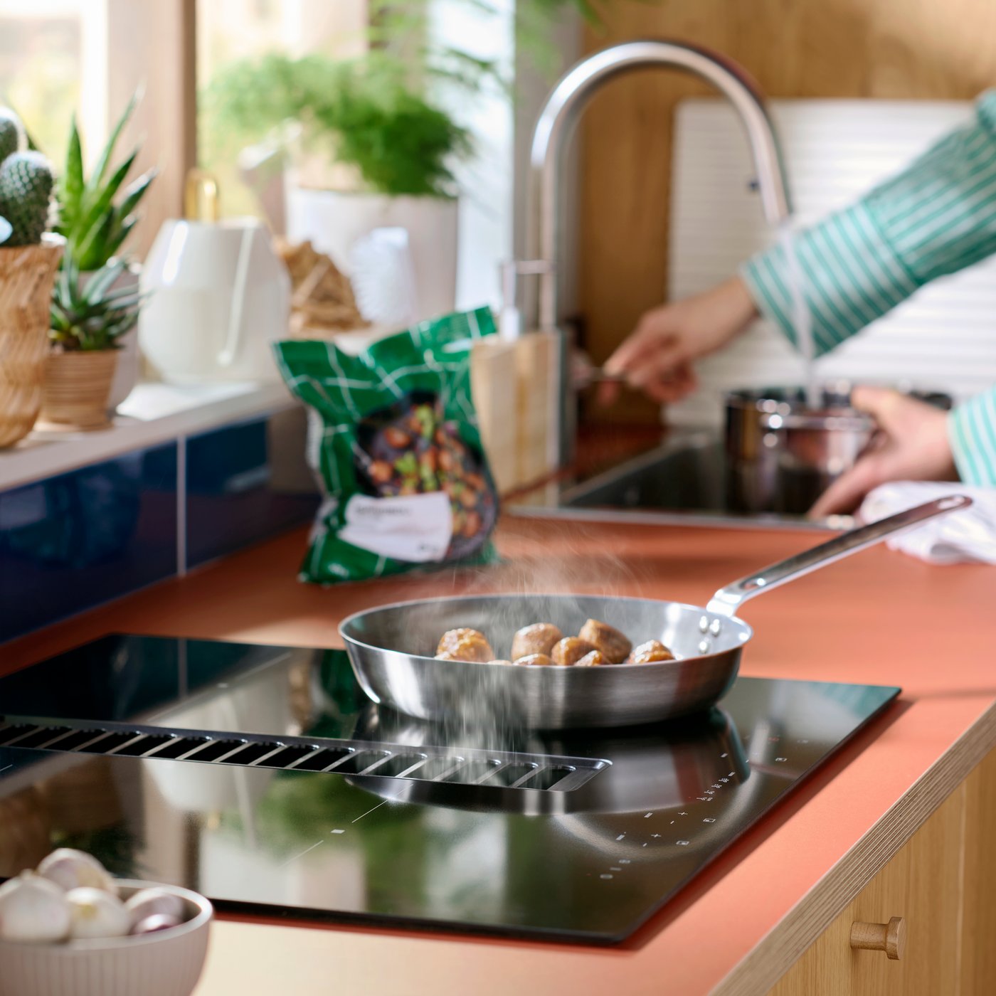 A frying pan is standing on a TÄCKNAN induction hob with integrated extractor and there’s a person in the background.